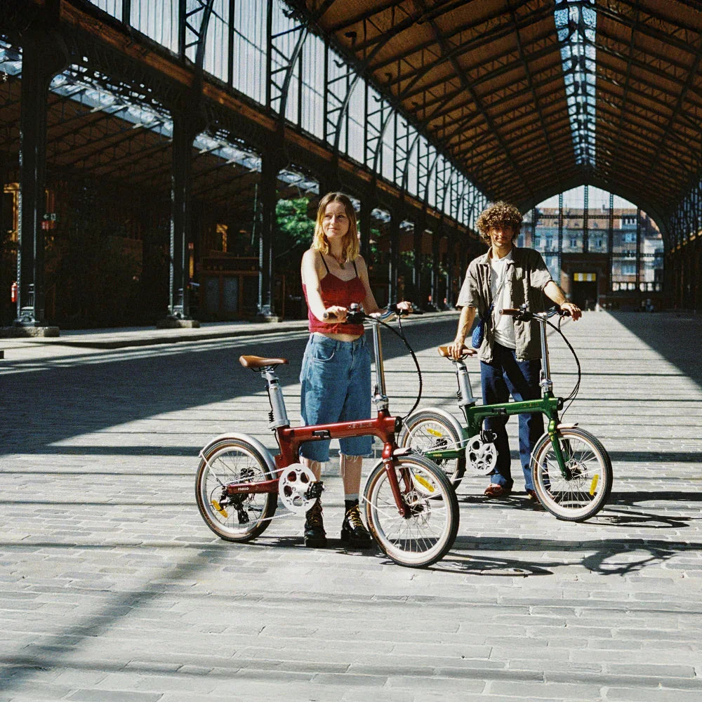 Two young adults standing with vintage-style folding bikes under a large industrial glass-roofed pavilion