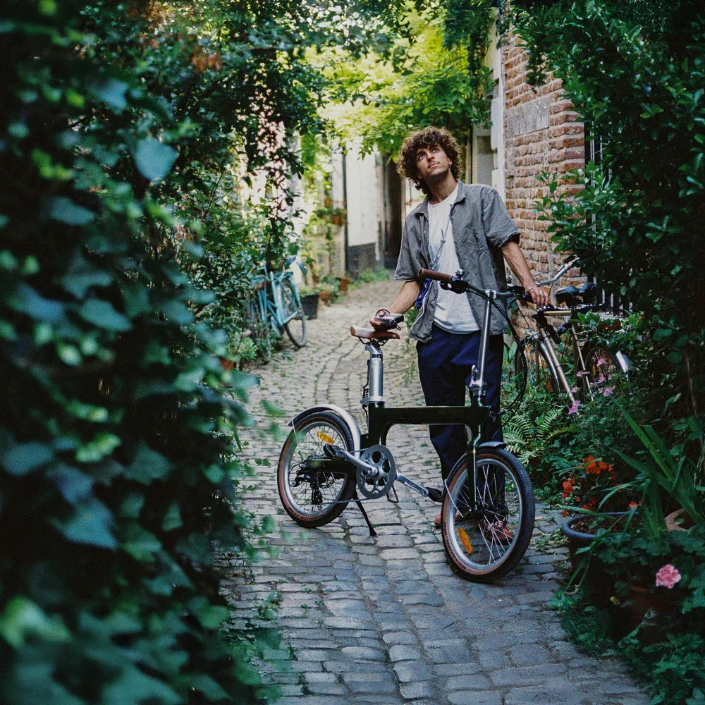 Young man with curly hair standing on a cobblestone alley holding a vintage-style electric bike surrounded by greenery and flowers