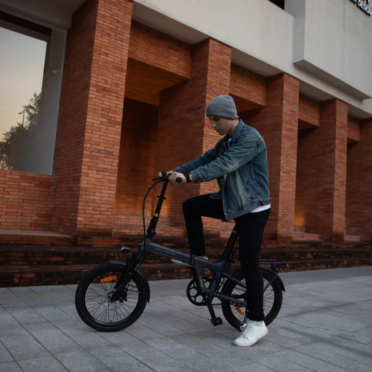 Man in denim jacket and gray beanie riding a black foldable electric bike in urban setting
