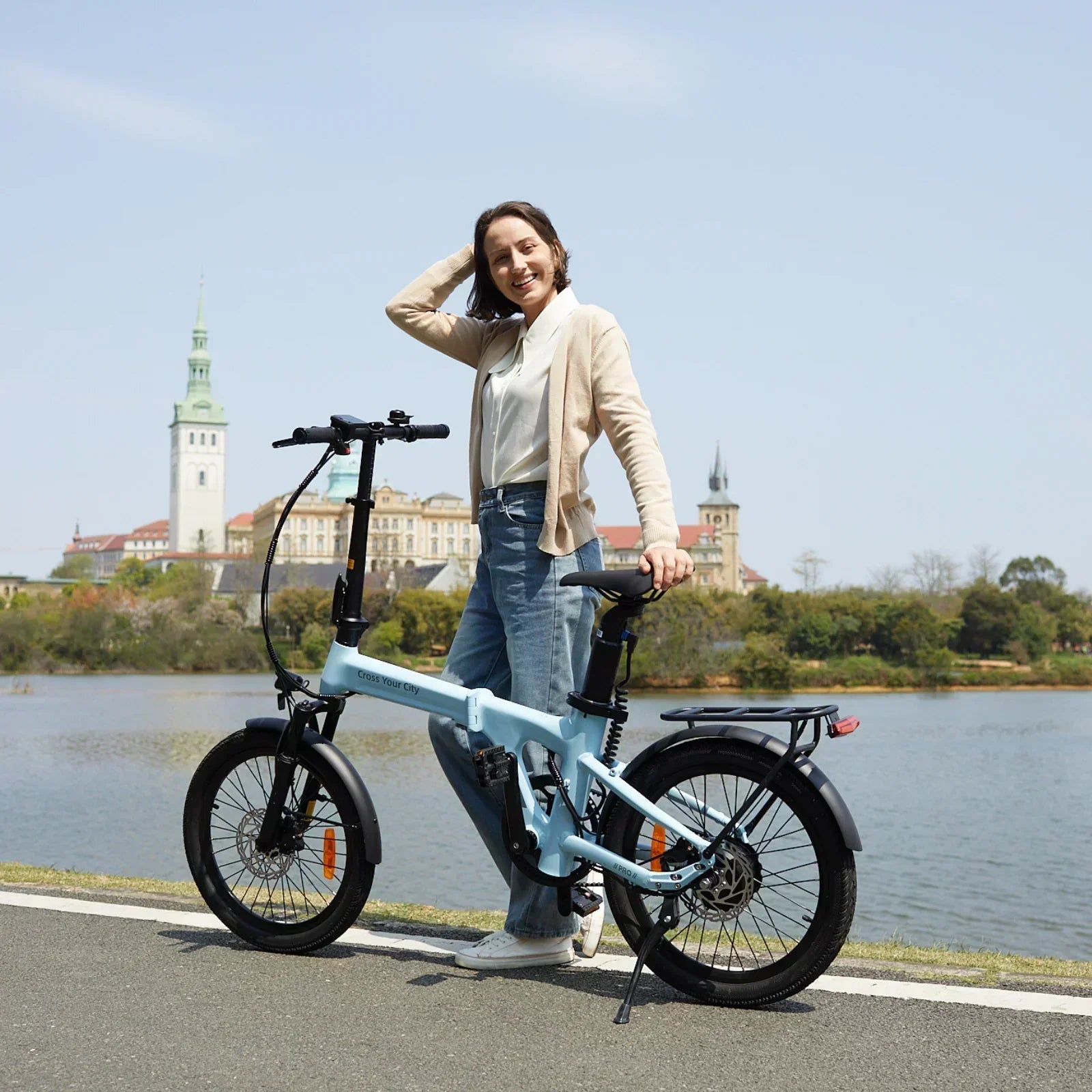 Woman standing beside light blue folding e-bike by a river with historic buildings in background