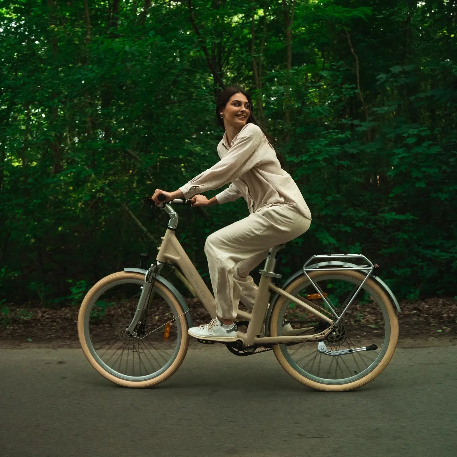 Woman riding beige electric bike on forest path, smiling in casual beige outfit and white sneakers