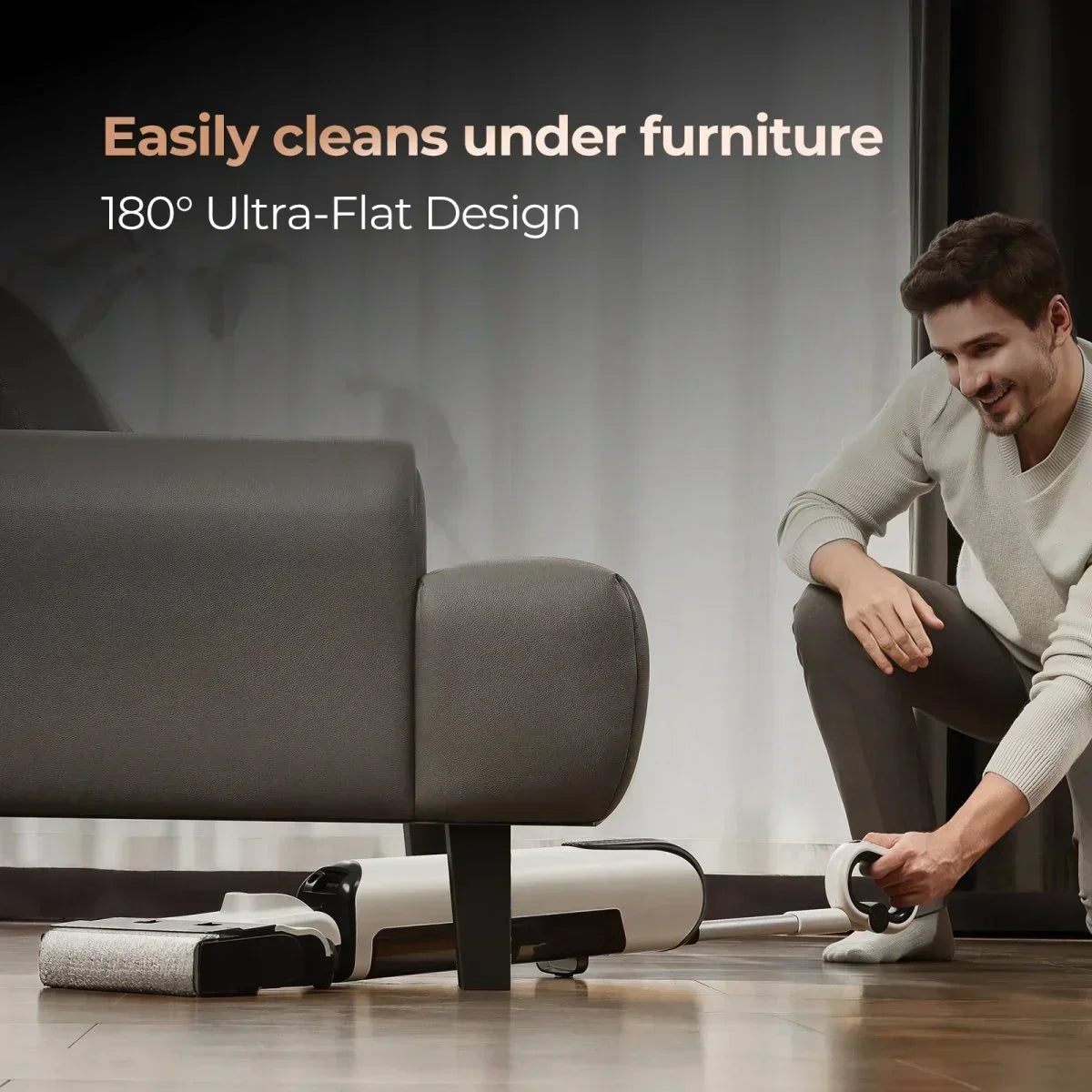 Man using ultra-flat vacuum cleaner to easily clean under gray sofa on wooden floor