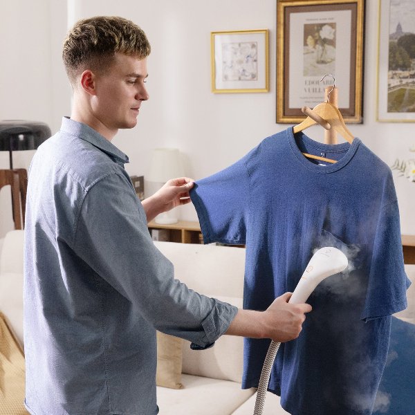 Man using handheld garment steamer to remove wrinkles from blue t-shirt in living room