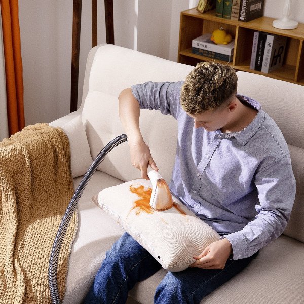Man using handheld steam cleaner to remove orange stain from beige cushion on white sofa indoors