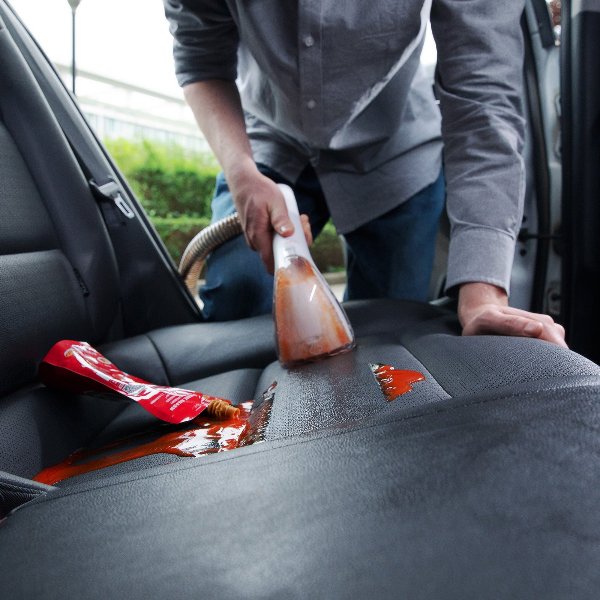 Person using steam cleaner to remove spilled ketchup from black car seat upholstery